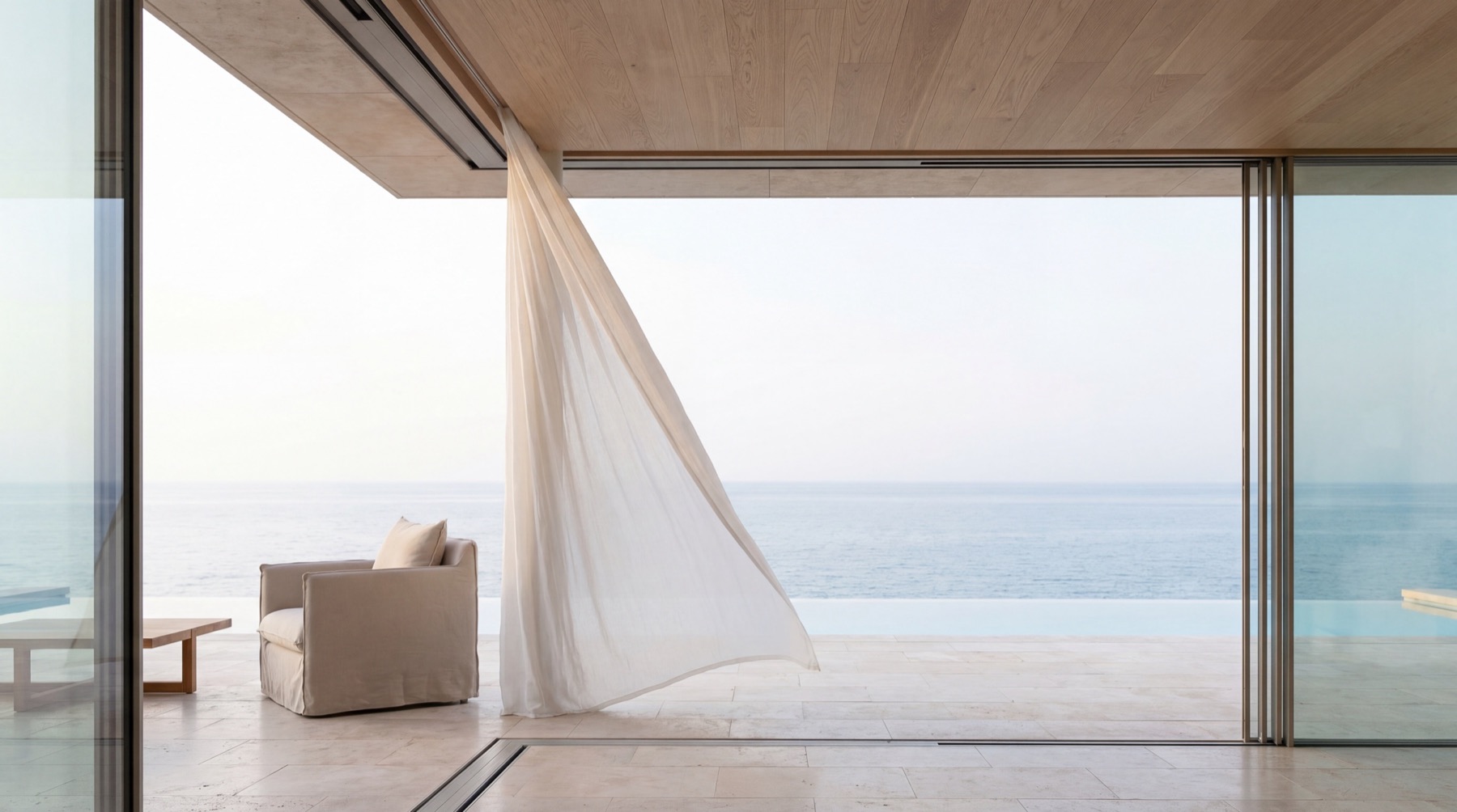 A sheer linen curtain lifting on the breeze through a fully opened glass wall, the sea beyond